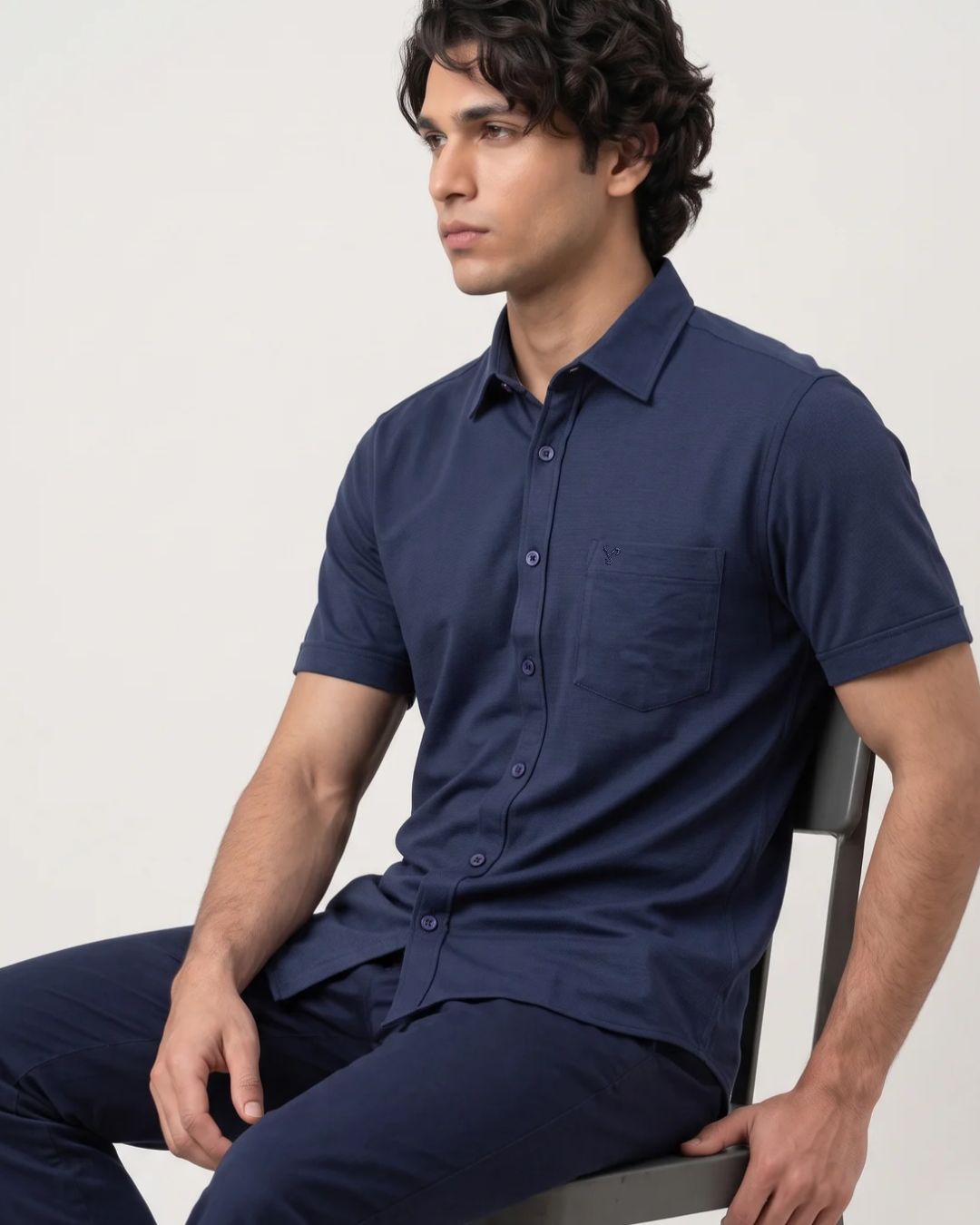 A man wearing a navy blue short-sleeve button-down shirt and matching navy pants, seated on a chair against a plain light background.