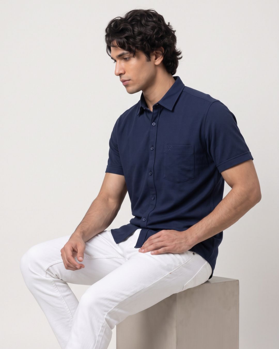 A man wearing a navy blue short-sleeve button-up shirt and white pants, sitting on a light gray concrete block against a plain off-white studio background.