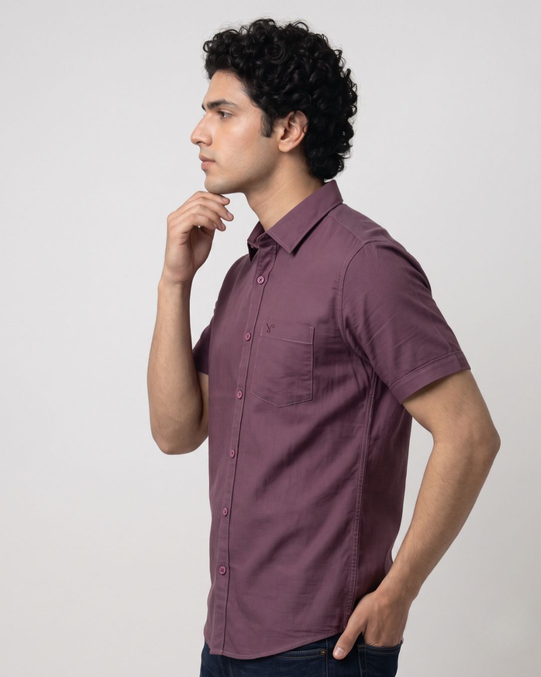 A man wearing a short-sleeve button-up shirt in a dusty plum purple color, standing in profile against a light gray studio background.