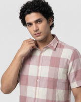 A man wearing a short-sleeve button-up shirt with a pink and off-white checkered pattern, posed against a light gray background.