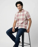 A man wearing a pink and white checkered short-sleeve button-up shirt and dark blue jeans, sitting on a black metal stool against a light gray studio background.
