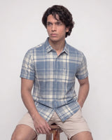 A man wearing a short-sleeve plaid button-up shirt in blue and cream, paired with beige shorts, sitting on a wooden stool against a plain light gray background.