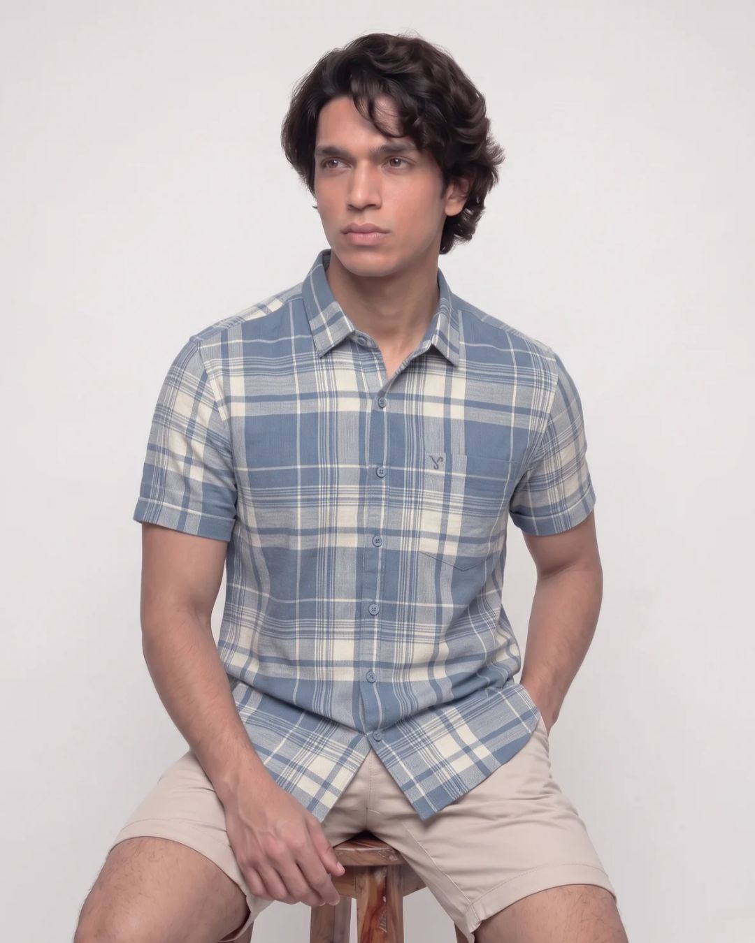 A man wearing a short-sleeve plaid button-up shirt in blue and cream, paired with beige shorts, sitting on a wooden stool against a plain light gray background.