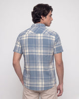 A man wearing a short-sleeve button-up plaid shirt in blue and white, paired with beige trousers, standing against a plain light gray background.
