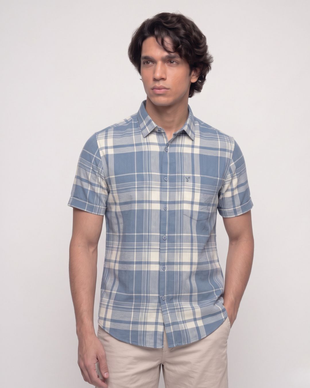 A man wearing a short-sleeve button-up plaid shirt in blue and white, paired with beige trousers, standing against a plain light gray background.