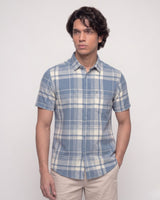 A man wearing a short-sleeve button-up plaid shirt in blue and white, paired with beige trousers, standing against a plain light gray background.