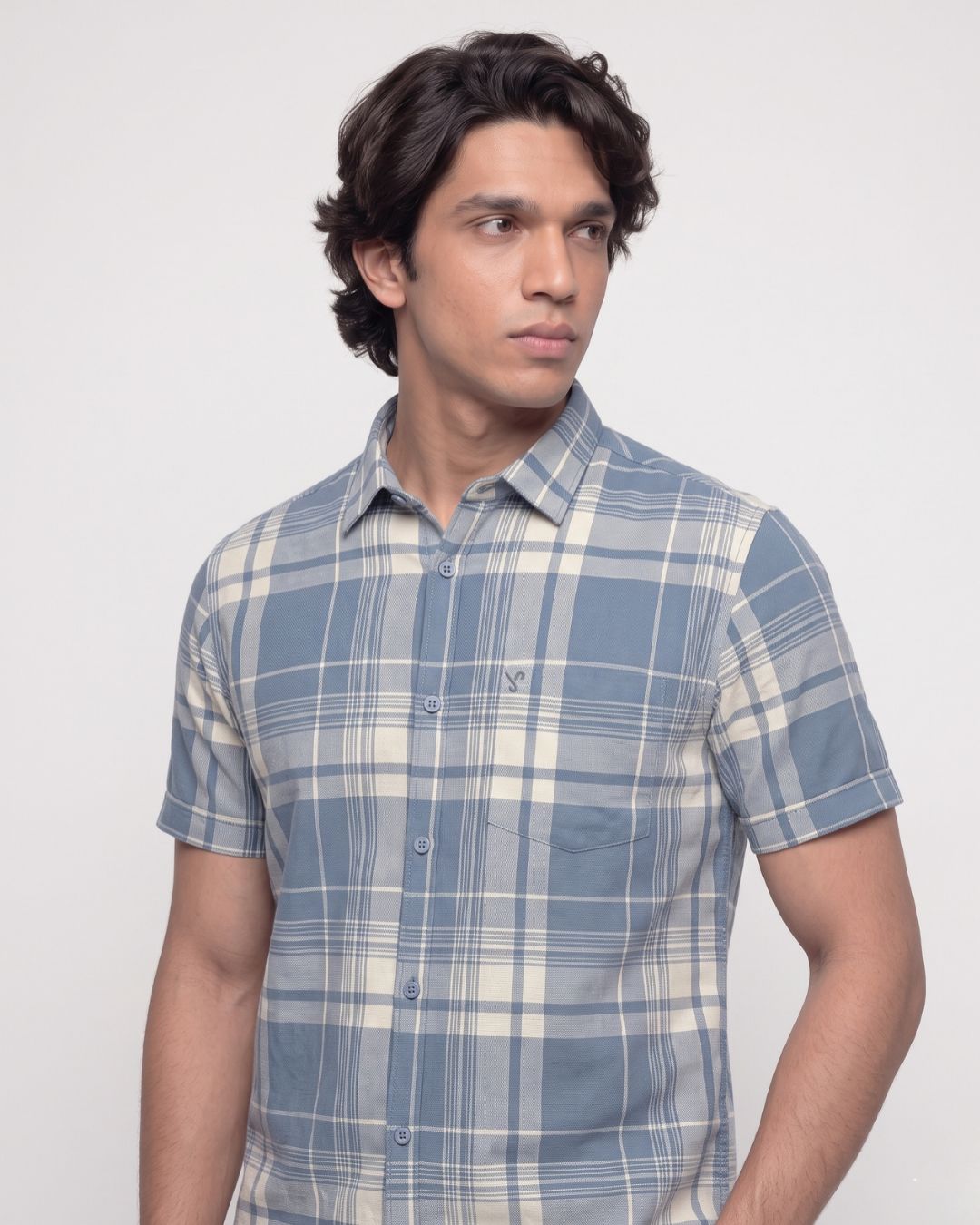 A man wearing a short-sleeve button-up plaid shirt in shades of blue and cream, photographed against a plain light gray studio background.