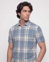 A man wearing a short-sleeve button-up plaid shirt in shades of blue and cream, photographed against a plain light gray studio background.