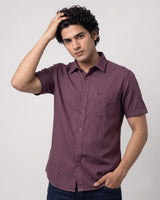 A man wearing a short-sleeve button-up shirt in a dusty plum purple color, paired with dark blue jeans, posing against a light gray studio background.
