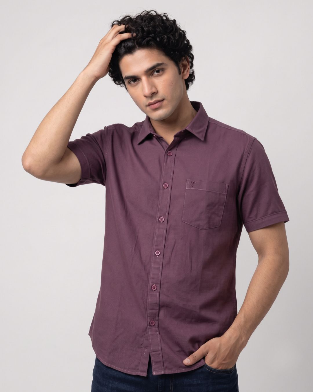 A man wearing a short-sleeve button-up shirt in a dusty plum purple color, paired with dark blue jeans, posing against a light gray studio background.