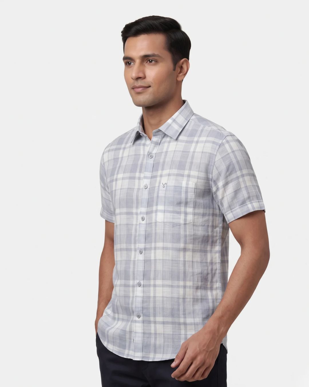 A man wearing a light gray and white plaid short-sleeve button-down shirt, standing against a solid light gray background.