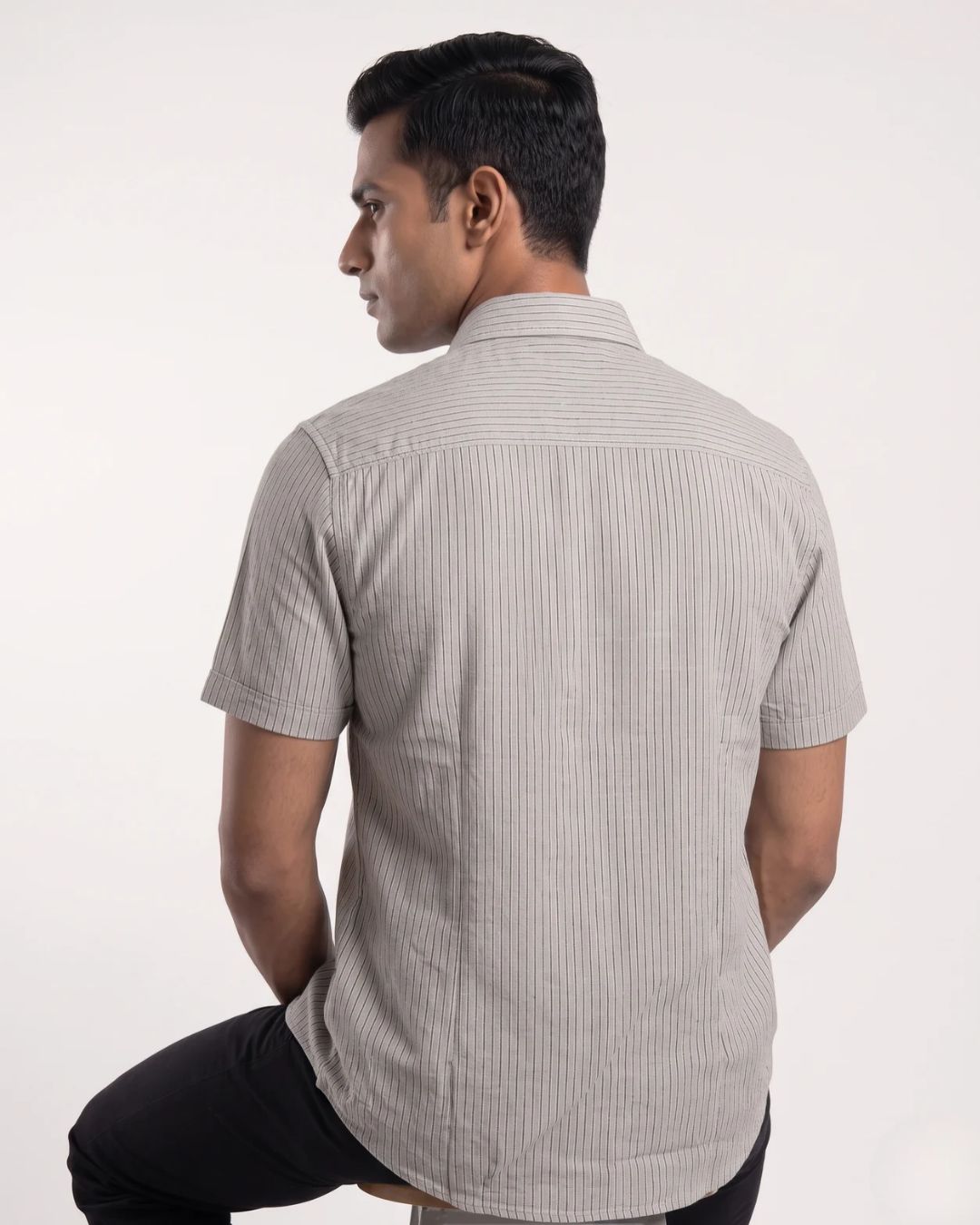 A man wearing a light gray and white vertically striped short-sleeve button-up shirt, shown from the back against a plain white studio background.