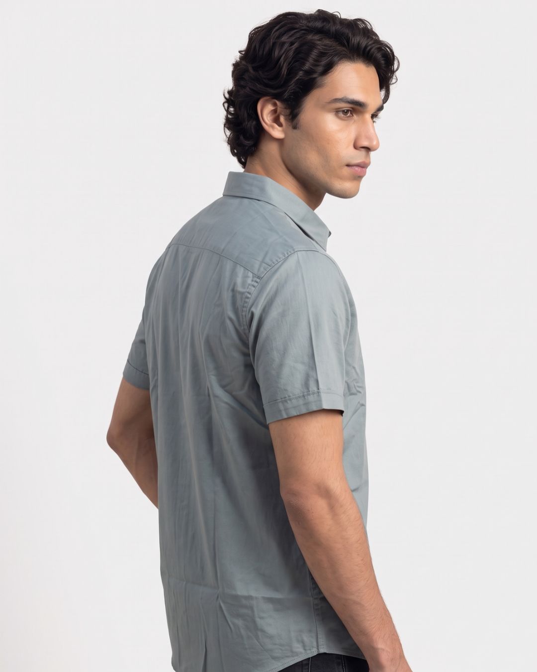 A man wearing a light gray short-sleeve button-up shirt, shown from a side-back angle against a plain light gray studio background.