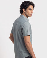 A man wearing a light gray short-sleeve button-up shirt, shown from a side-back angle against a plain light gray studio background.