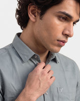 A close-up of a man wearing a light gray button-down shirt, adjusting the collar with his hand against a plain light gray background.