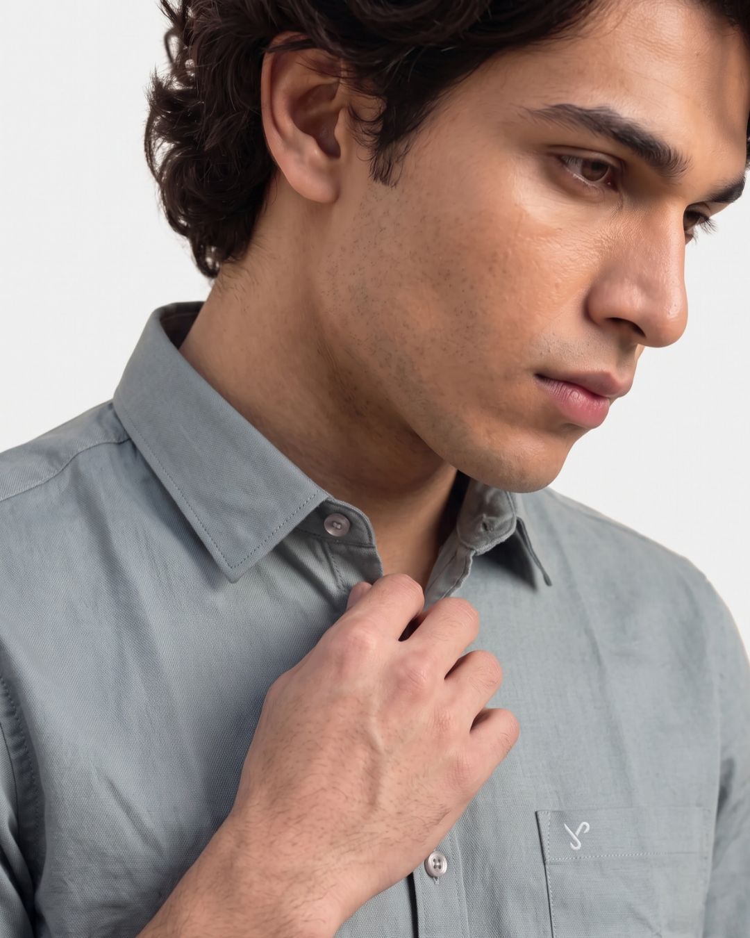 A close-up of a man wearing a light gray button-down shirt, adjusting the collar with his hand against a plain light gray background.