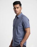 A man wearing a slate blue short-sleeve button-up shirt, photographed from the waist up against a plain light gray studio background.