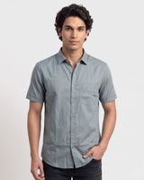 A man wearing a light gray short-sleeve button-down shirt with a chest pocket, paired with dark jeans, standing against a plain light gray background.