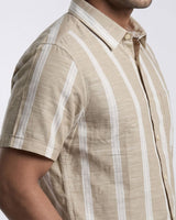 A man wearing a beige and white vertically striped short-sleeve button-up shirt, sitting on a stool against a light gray studio background.