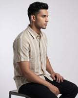 A man wearing a beige and white vertically striped short-sleeve button-up shirt, sitting on a stool against a light gray studio background.