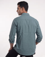A man wearing a muted blue-gray long-sleeve button-down shirt with black trousers, standing against a plain light gray studio background.