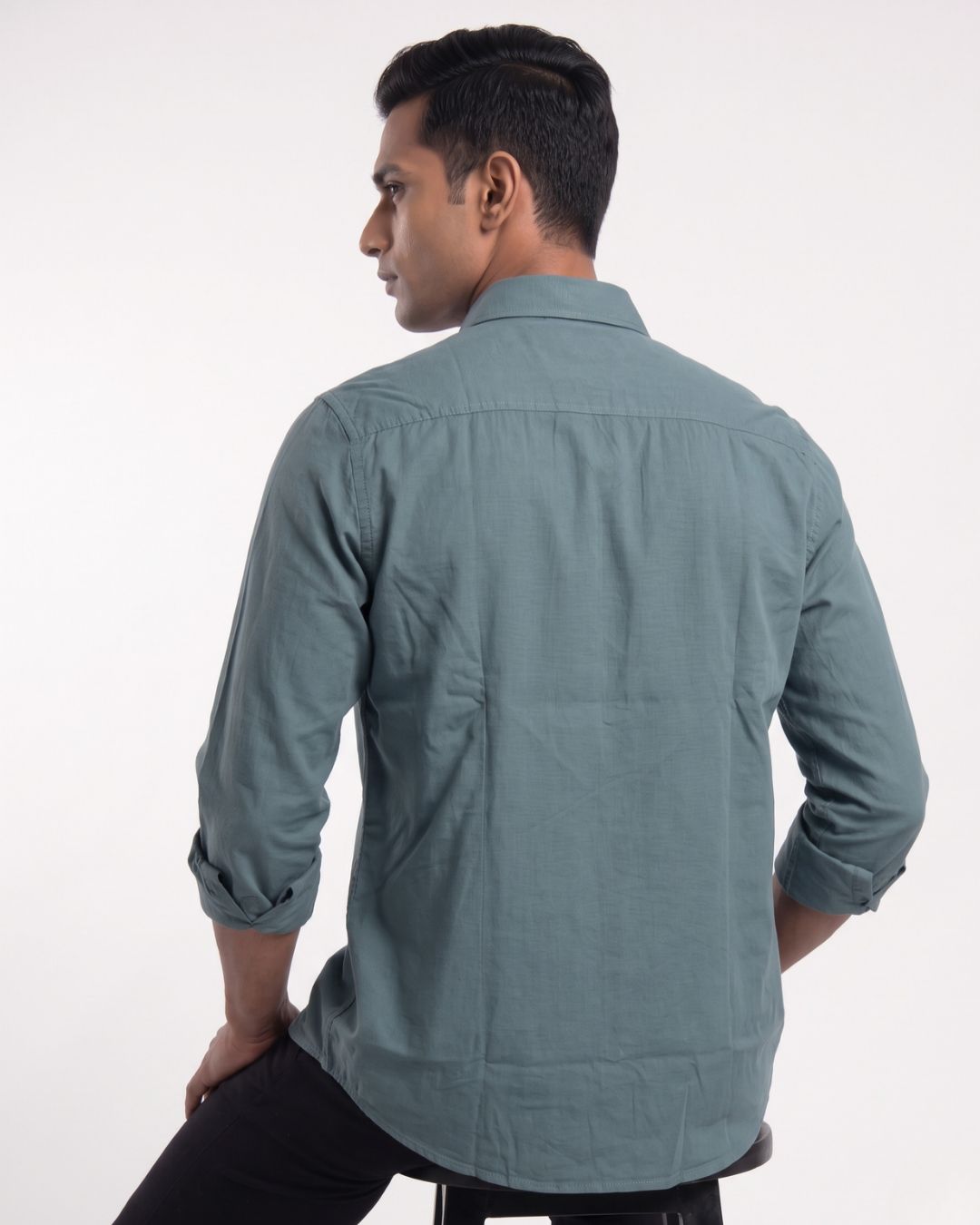 A man wearing a muted blue-gray long-sleeve button-down shirt with black trousers, standing against a plain light gray studio background.