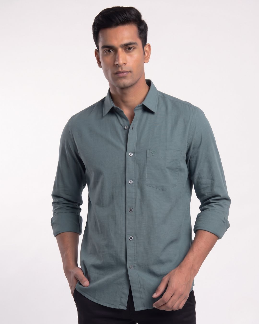 A man wearing a muted blue-gray long-sleeve button-down shirt with black trousers, standing against a plain light gray studio background.