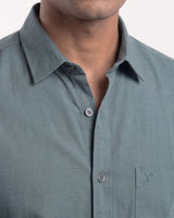 A man wearing a muted blue-gray long-sleeve button-down shirt with black trousers, standing against a plain light gray studio background.