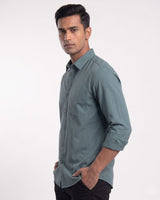 A man wearing a muted blue-gray long-sleeve button-down shirt with black trousers, standing against a plain light gray studio background.