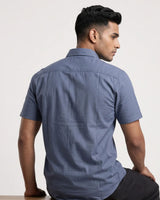 A man wearing a slate blue short-sleeve button-up shirt, shown from the back and side profile against a plain off-white studio background.