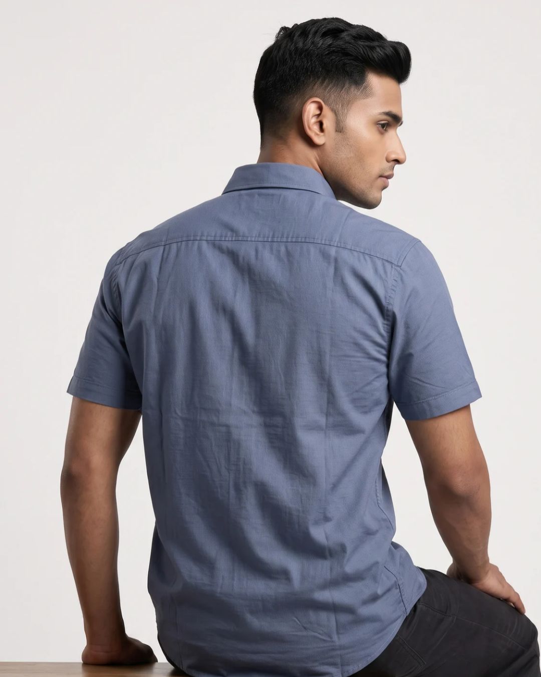 A man wearing a slate blue short-sleeve button-up shirt, shown from the back and side profile against a plain off-white studio background.