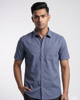 A man wearing a short-sleeve button-down shirt in slate blue, photographed against a light gray studio background.