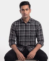 A man wearing a long-sleeve black and gray plaid button-down shirt, sitting against a plain light gray background.