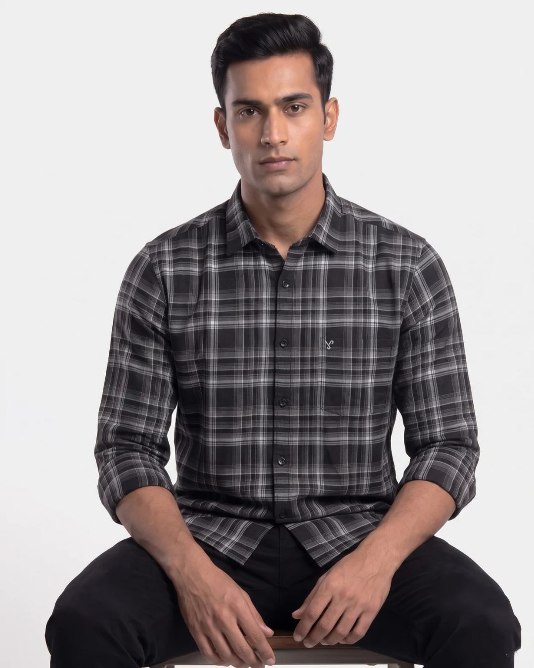A man wearing a long-sleeve black and gray plaid button-down shirt, sitting against a plain light gray background.