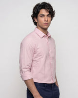 A man wearing a light pink long-sleeve button-down shirt with dark navy trousers, standing against a plain light gray studio background.