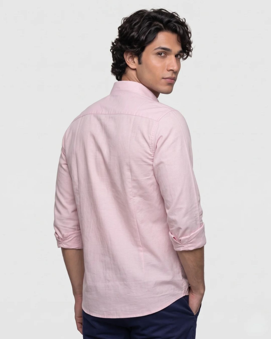 A man wearing a light pink long-sleeve linen shirt with rolled-up sleeves, shown from a three-quarters back view against a plain light gray background.