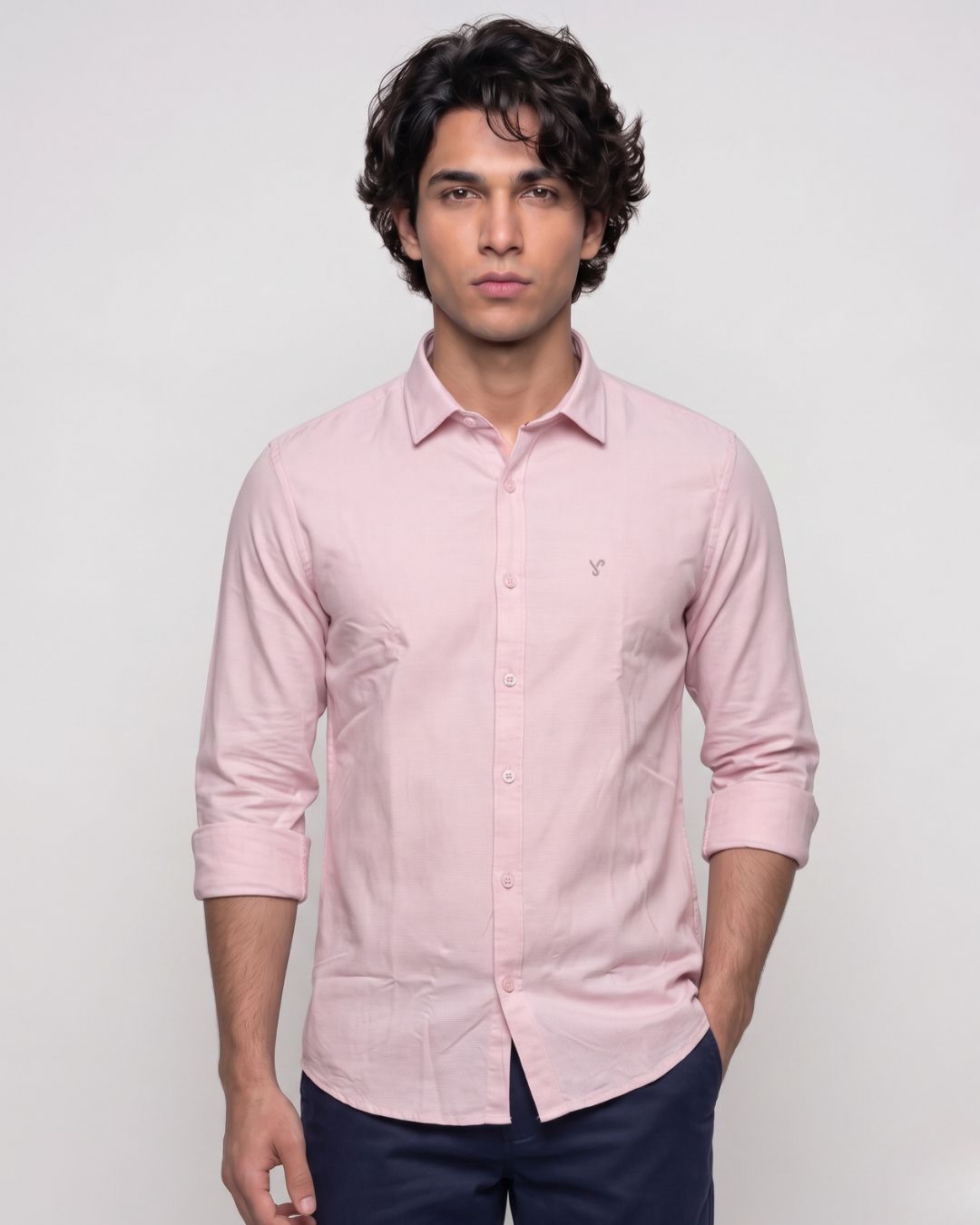 A man wearing a light pink long-sleeve button-down shirt with rolled-up sleeves and dark navy trousers, standing against a light gray studio background.