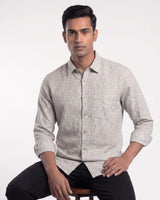 A man wearing a light beige long-sleeve button-up shirt with a fine white micro-pattern, paired with black trousers, seated against a plain white studio background.