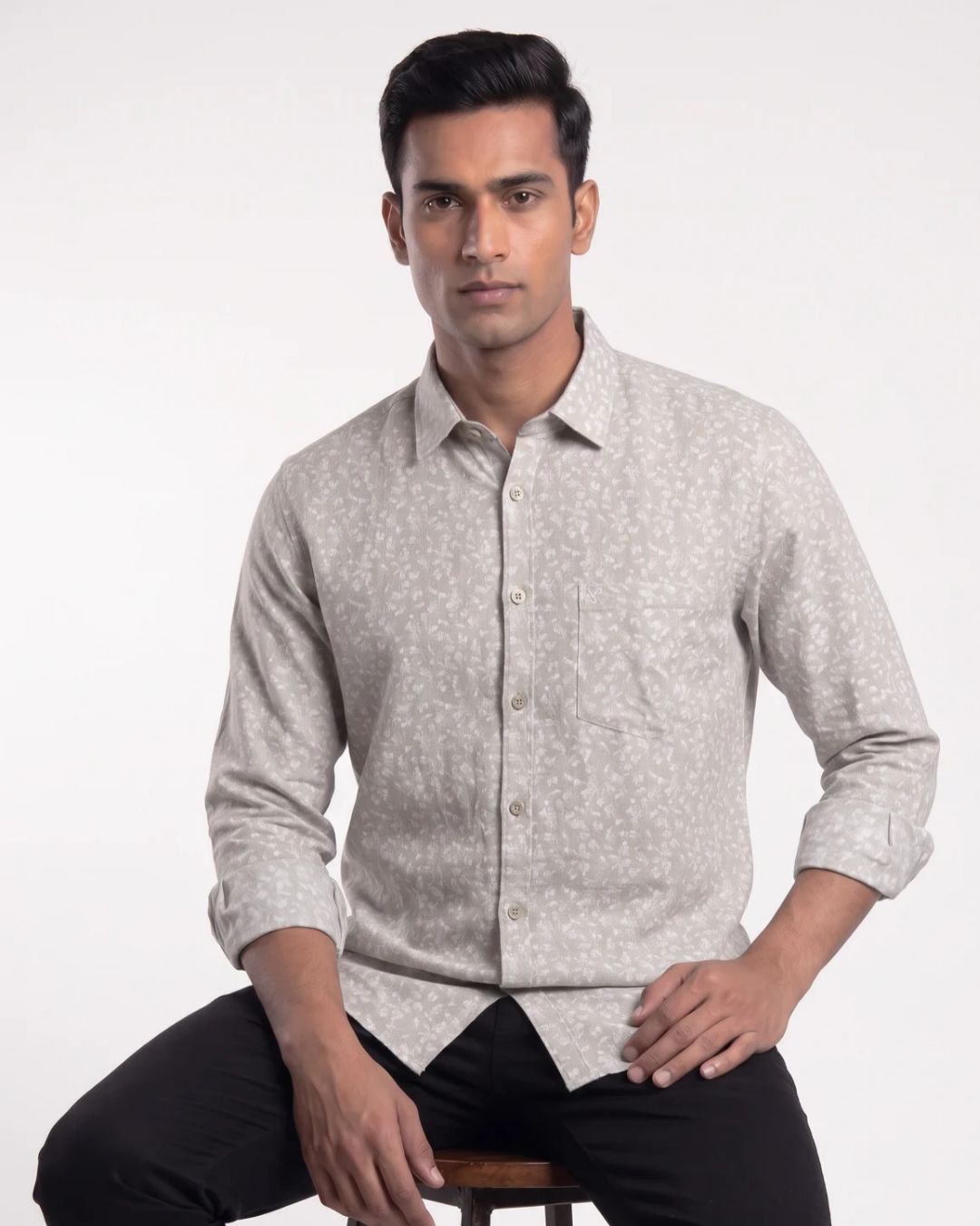 A man wearing a light beige long-sleeve button-up shirt with a fine white micro-pattern, paired with black trousers, seated against a plain white studio background.