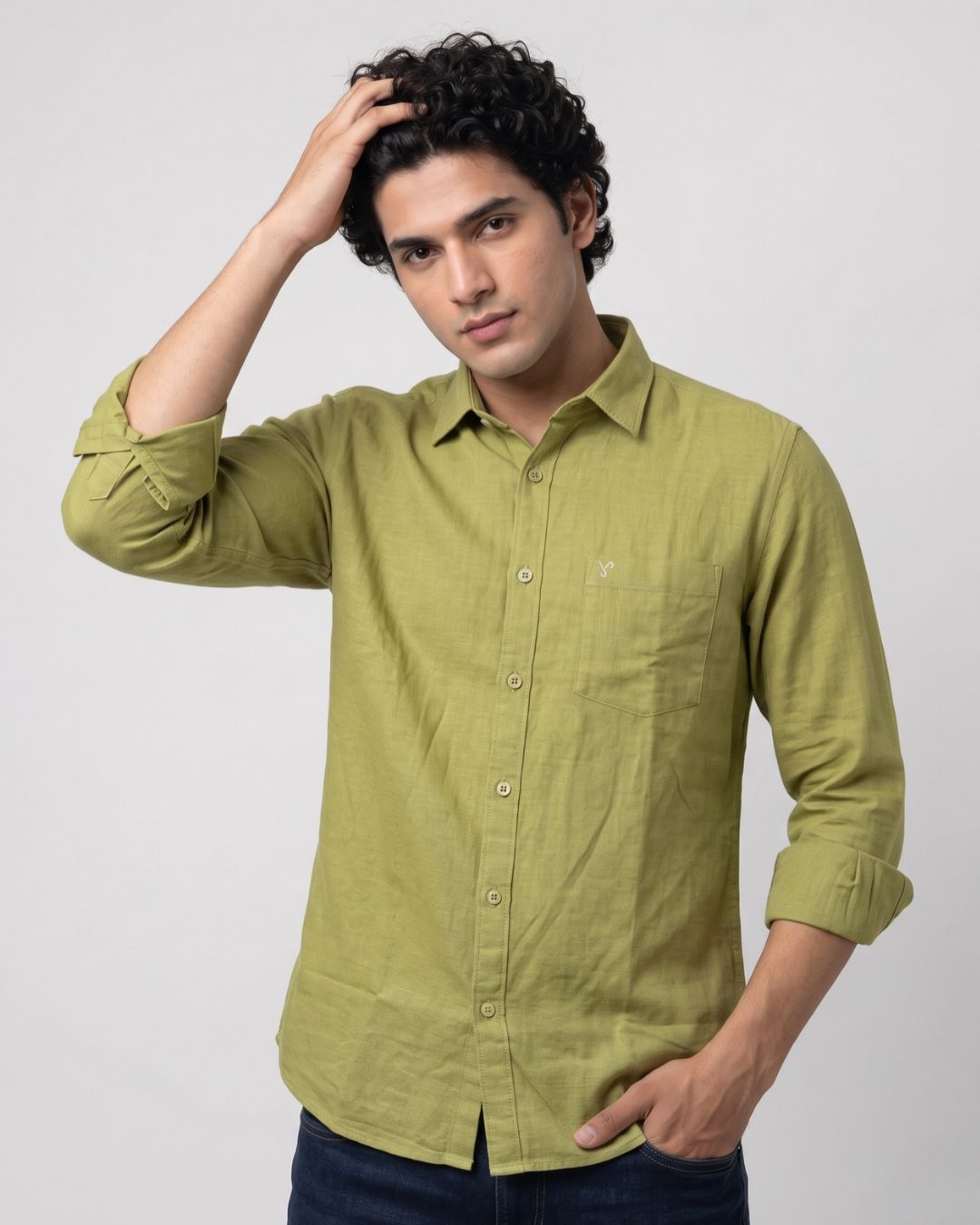 A man wearing a light olive green linen button-down shirt with dark blue jeans, posing against a light gray studio background.