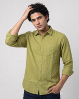 A man wearing a light olive green linen button-down shirt with dark blue jeans, posing against a light gray studio background.