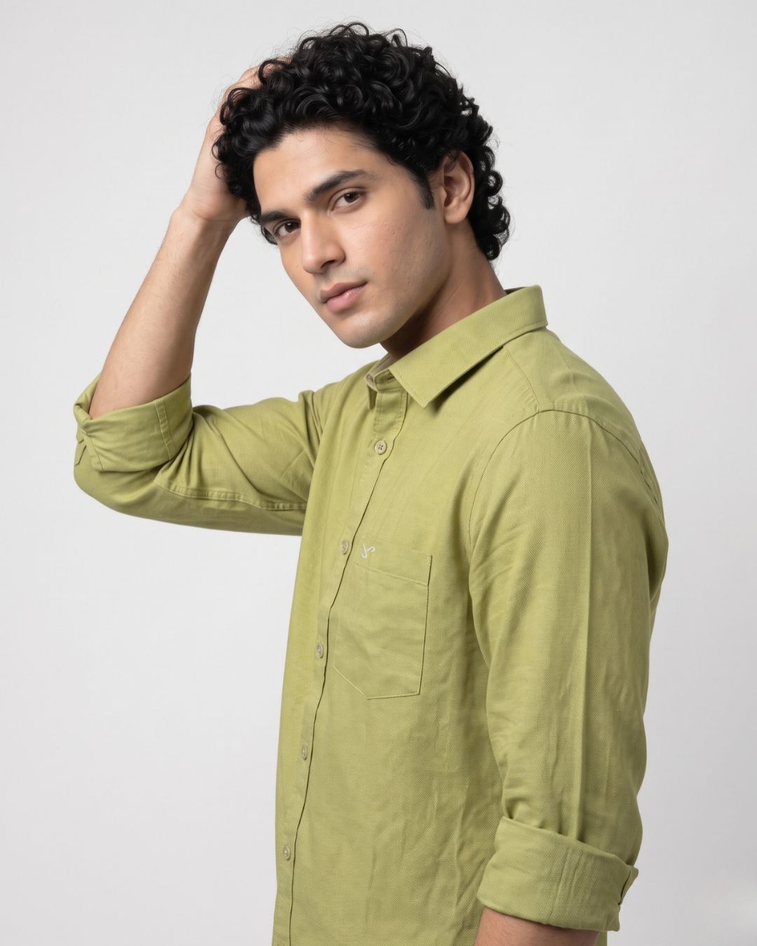 A man with curly black hair wearing a light olive green long-sleeve button-up shirt, posing against a light gray studio background.