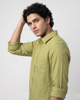 A man with curly black hair wearing a light olive green long-sleeve button-up shirt, posing against a light gray studio background.