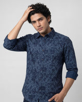 A man wearing a dark blue patterned long-sleeve button-up shirt, posing against a light gray studio background.