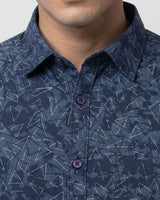 A man wearing a dark blue patterned long-sleeve button-up shirt, posing against a light gray studio background.