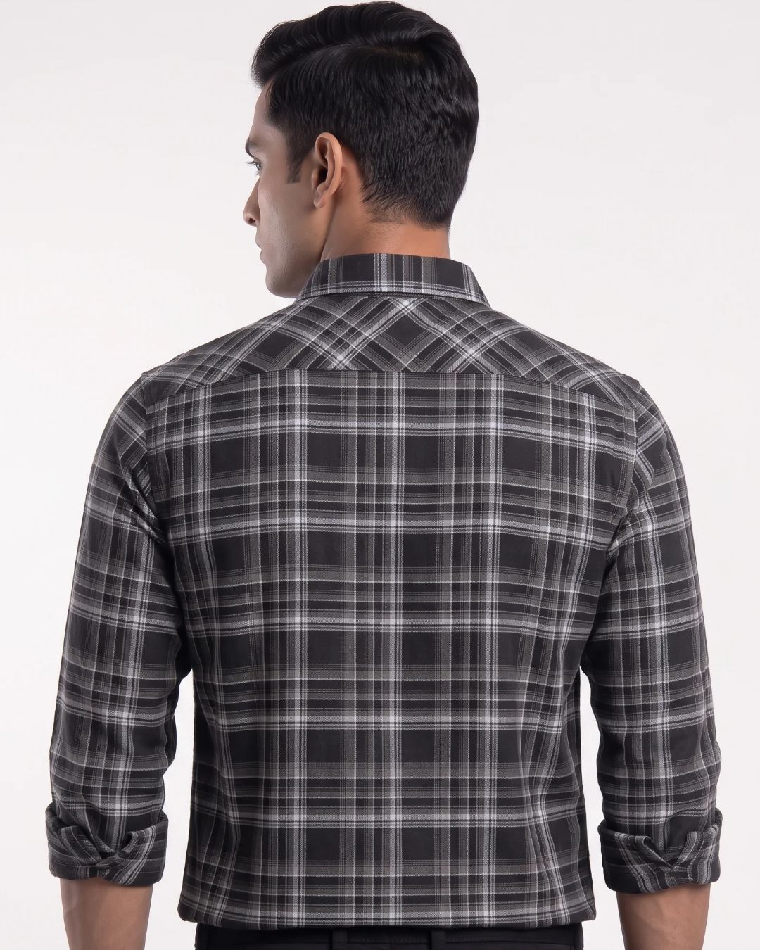 A man viewed from the back wearing a long-sleeve plaid button-up shirt in black, gray, and white, with the sleeves rolled up against a plain light background.