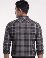 A man viewed from the back wearing a long-sleeve plaid button-up shirt in black, gray, and white, with the sleeves rolled up against a plain light background.