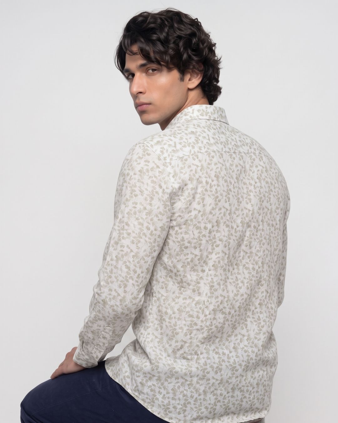 A man wearing a light beige long-sleeve button-up shirt with a small gray abstract pattern, shown from a three-quarters back view against a plain light gray studio background.