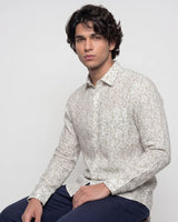 A man wearing a light beige long-sleeve button-up shirt with a small, dense abstract print, paired with navy blue pants, seated against a plain light gray background.