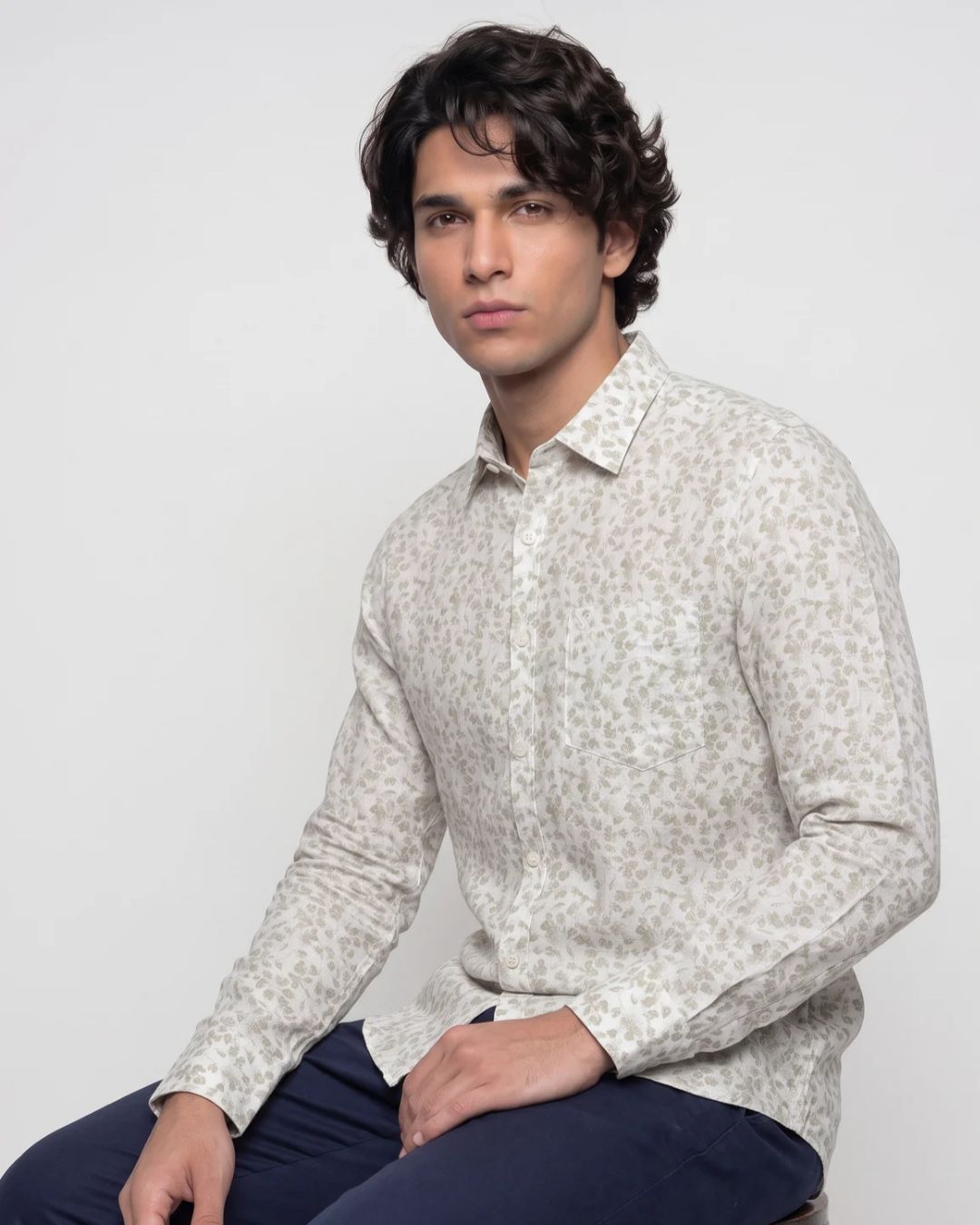 A man wearing a light beige long-sleeve button-up shirt with a small, dense abstract print, paired with navy blue pants, seated against a plain light gray background.